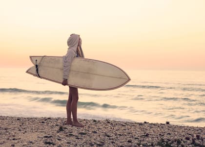 A person in a hoodie holding a surfboard on a beach. at Seacoast Residences Apartments, Oakland, ME, 03904