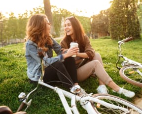 Two Women Sitting In Grass Having Conversation Next to Their Bikes 