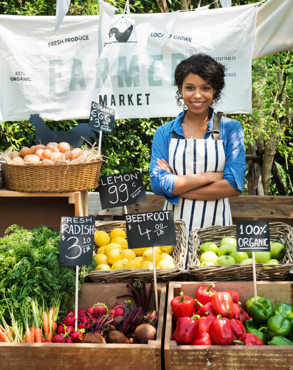 a woman standing in front of a vegetable stand at a farmers market