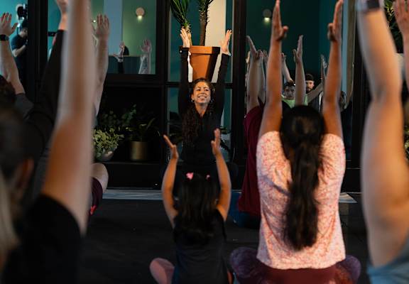 A group of people are sitting on the floor with their hands raised in a yoga class.
