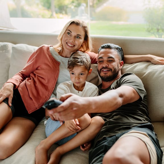 A family of three is sitting on a couch, with the man holding a remote control.
