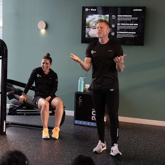 A man and a woman are in a gym, the man is standing and talking while the woman is sitting on a machine.
