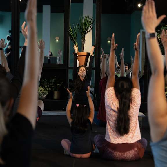 A group of people are sitting on the floor with their hands raised in a yoga class.