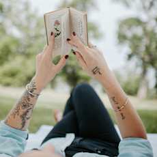 A person with tattoos on their arms is reading a book outdoors.