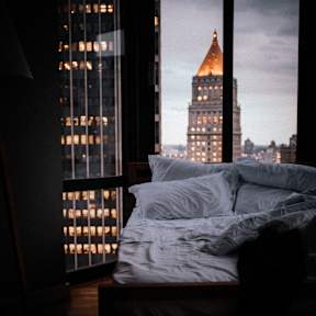 A bed with white sheets sits in front of a window with a view of a cityscape.