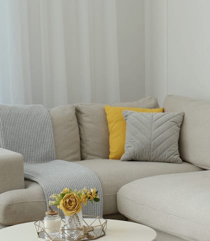 A grey couch with a yellow pillow and a small table with a vase of flowers on it.