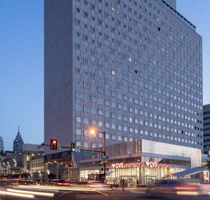 A city street at dusk with cars passing by and buildings in the background.