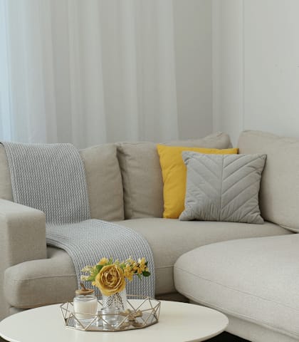 A grey couch with a yellow pillow and a small table with a vase of flowers on it.