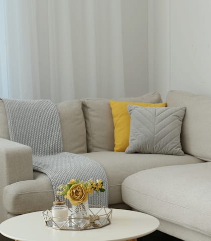 A grey couch with a yellow pillow and a small table with a vase of flowers on it.
