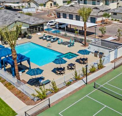 A tennis court is surrounded by a pool and palm trees.