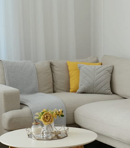 A grey couch with a yellow pillow and a small table with a vase of flowers on it.