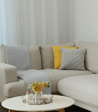 A grey couch with a yellow pillow and a small table with a vase of flowers on it.