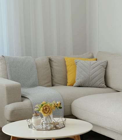 A grey couch with a yellow pillow and a small table with a vase of flowers on it.