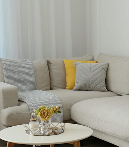 A grey couch with a yellow pillow and a small table with a vase of flowers on it.