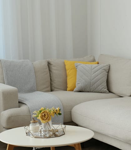 A grey couch with a yellow pillow and a small table with a vase of flowers on it.