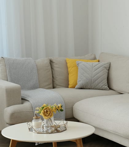 A grey couch with a yellow pillow and a small table with a vase of flowers on it.