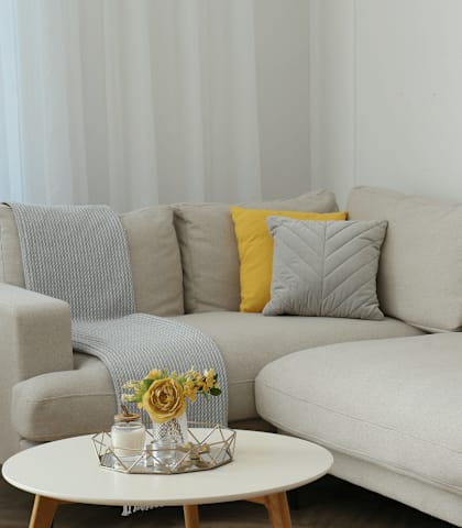 A grey couch with a yellow pillow and a small table with a vase of flowers on it.