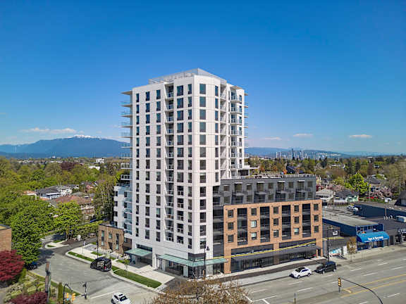 A tall white building with a balcony on the top floor is surrounded by other buildings and trees.