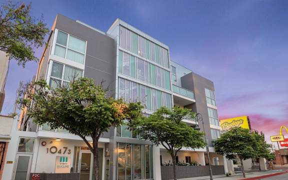 A modern building with a glass facade and a sign that reads "1047s" is surrounded by trees and a sidewalk.