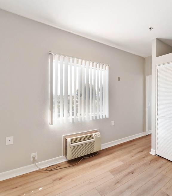 A room with a window covered by blinds and a wall-mounted air conditioner. at Legacy Hills at Poway 55+ Apartments, Poway, California