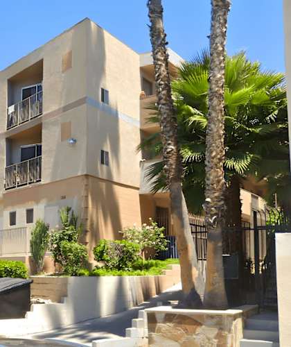 A modern building with a balcony is surrounded by trees and parked cars.