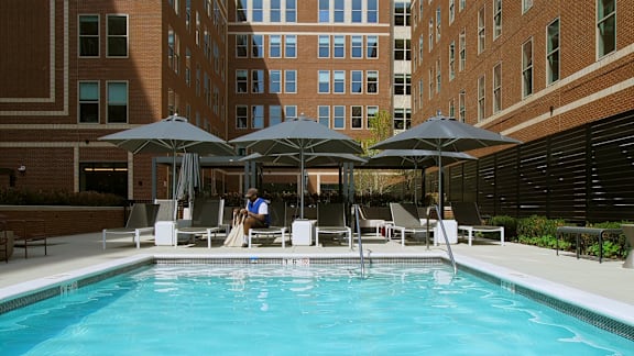 A man is sitting on a chair under an umbrella by a pool.