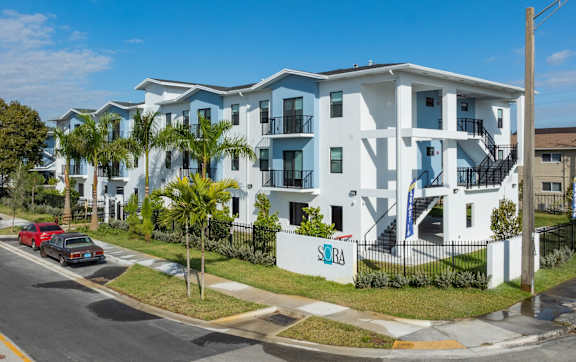 An image of Sora Apartments building and the front sign. Green grass and palm trees in the front.