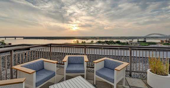 Two chairs and a table on a balcony overlooking a river at sunset.