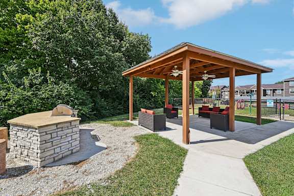 A wooden pavilion with a stone structure and a fire pit in the middle of a grassy area.