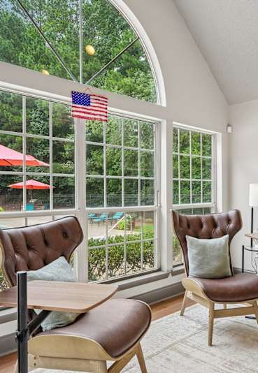 A room with a chair and a table in front of a window with a view of a pool and trees.