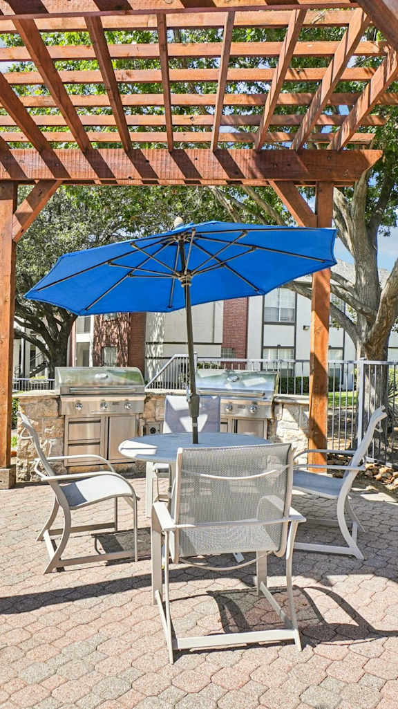 A wooden pergola with a blue umbrella is in the foreground of a brick patio.