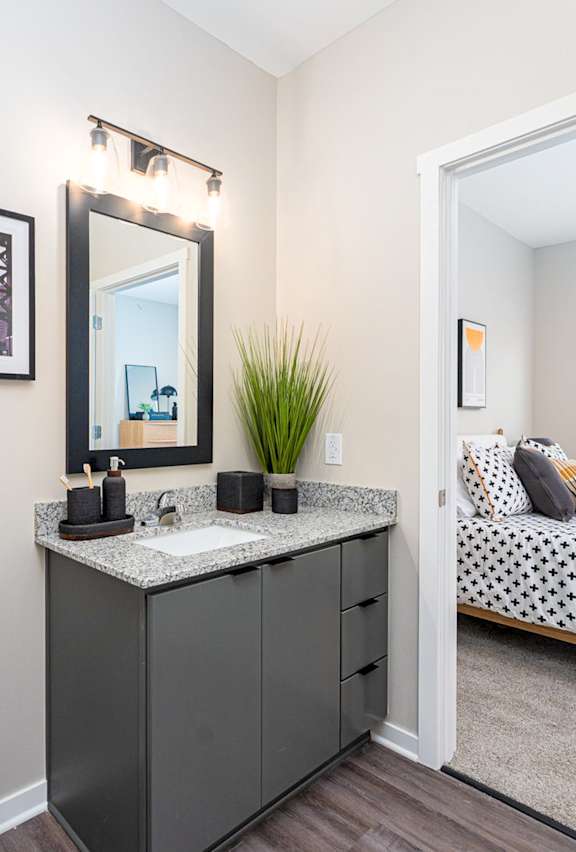 A bathroom with a toilet, sink, mirror, and a bed in the background.