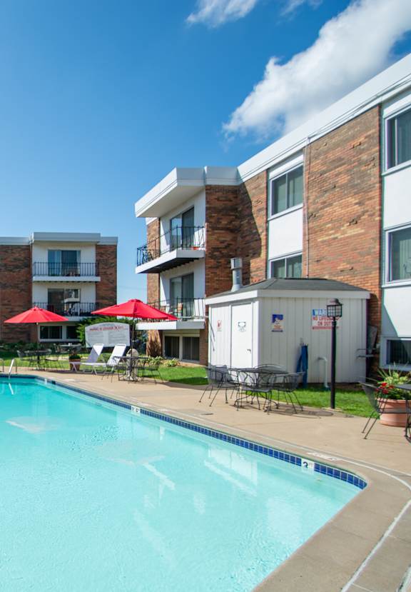 A swimming pool surrounded by red umbrellas and lounge chairs in front of apartment buildings.