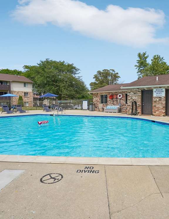 Outdoor swimming pool with expansive sundeck with lounge chairs. Exterior of Brighton Cove can be seen in the background. A pool house is off to the right. 