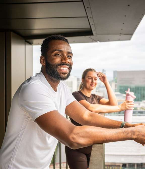 A man and a woman are standing on a balcony with a cityscape in the background.