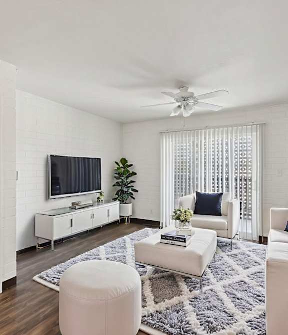A living room with a white couch, a white chair, a white ottoman, a white rug, a white ceiling fan, a white door, a white cabinet, a white television, and a white wall.