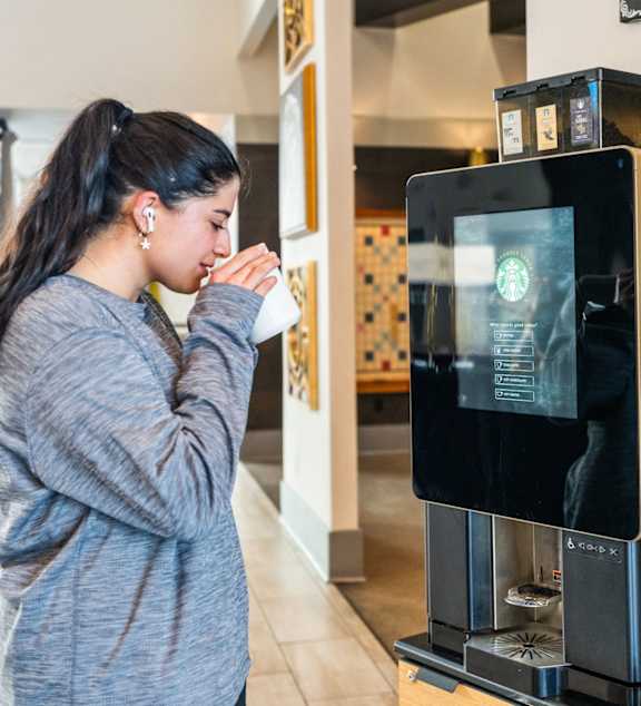 A woman is drinking from a Starbucks cup while standing in front of a Starbucks vending machine.