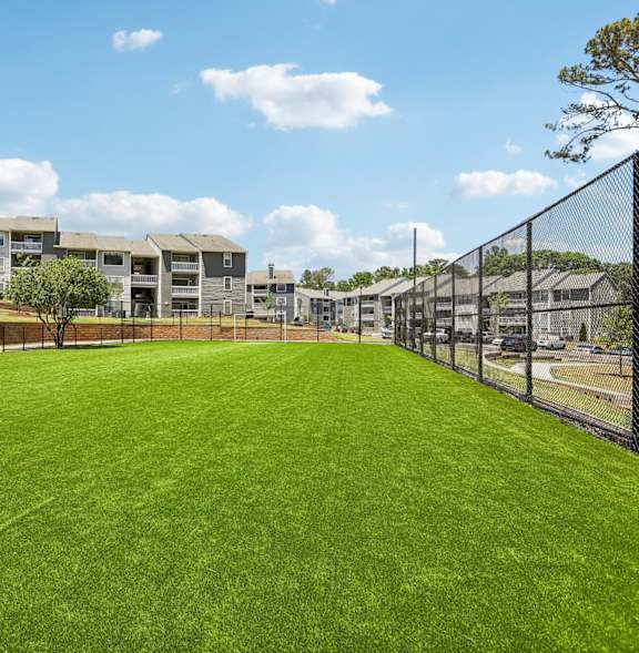 A large grassy field with a fence and apartment buildings in the background.