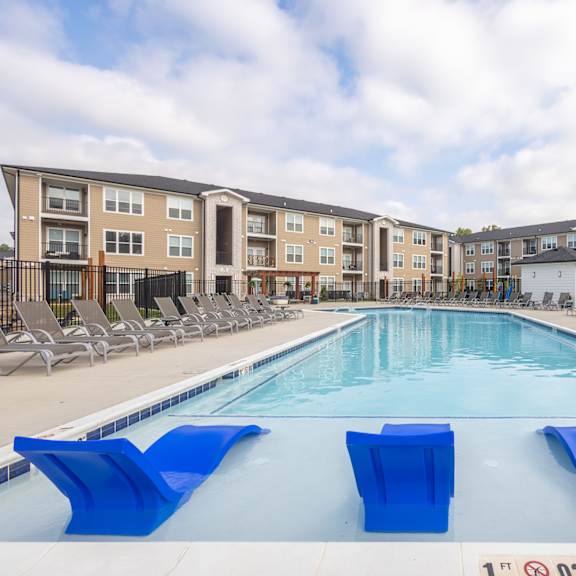 A swimming pool with blue lounge chairs and apartment buildings in the background.