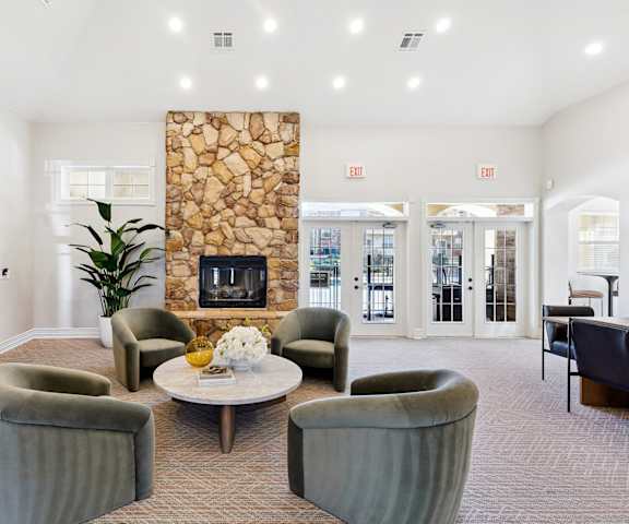 A large, well-lit resident lounge with wooden accents, windows, a door to the outside, decorations, a plush couch, bookshelves, tables, chairs, a TV, and a fireplace at Quail Landing Apartment Homes. 
