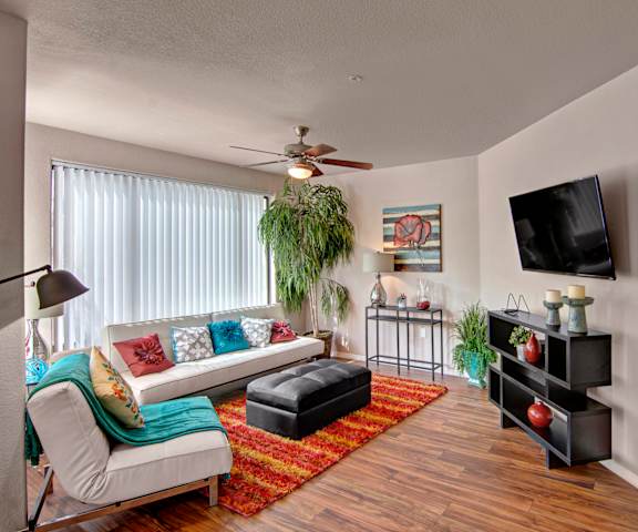 The spacious living room with a white couch, turquoise and pink throw pillows, large windows, wall art, plush chair, coffee table, decorations, overhead lighting with a fan, a red, orange, and yellow rug, large windows, a TV, and glass doors to the balcony at the Hawthorne Apartment Homes. 