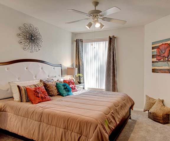 A spacious bedroom with a queen-size bed with a tan bedspread, a dresser, floral wall art, lamps, decorations, a large window with curtains, and nightstands with lamps at the Hawthorne Apartment Homes. 