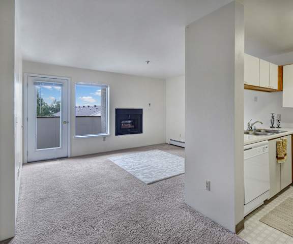 A view of the empty carpeted living room with a window, a fireplace, a door to the balcony, a rug, and the kitchen with white appliances and large, white cabinets at Silver Ridge Apartment Homes. 