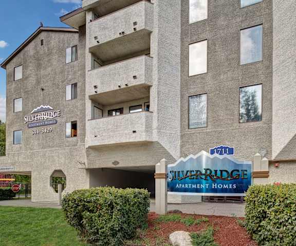 The blue and silver exterior sign surrounded by bushes with the gray apartment building in the background at Silver Ridge Apartment Homes. 
