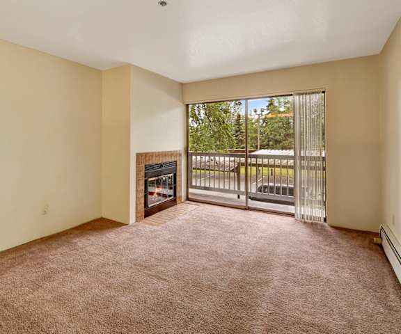 A large, empty living room with carpeting, a fireplace, and a sliding glass door to the balcony at Susitna Ridge Apartment Homes. 