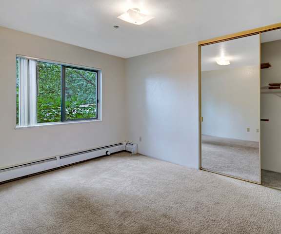 A large, empty carpeted bedroom with overhead lighting, a window showing the trees, and a closet at Susitna Ridge Apartment Homes. 