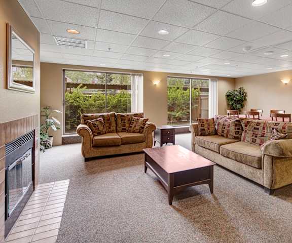 The amazing resident lounge with a fireplace with a mirror above it, couches, decorations, large windows with curtains, a coffee table, a dining table and chairs with a plaid tablecloth, and a plant in the corner at Susitna Ridge Apartment Homes. 