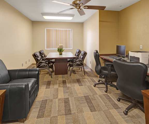 The business center with a desk with two monitors, two keyboards, and a printer, a leather couch, a meeting table with leather chairs surrounding it, overhead lighting, a ceiling fan, and a window with blinds at Susitna Apartment Homes. 