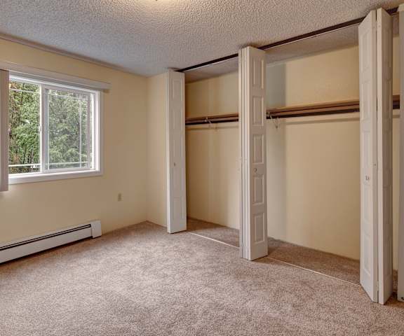A large, empty carpeted bedroom with overhead lighting, closets, a window, and a closet at Taiga Apartment Homes. 