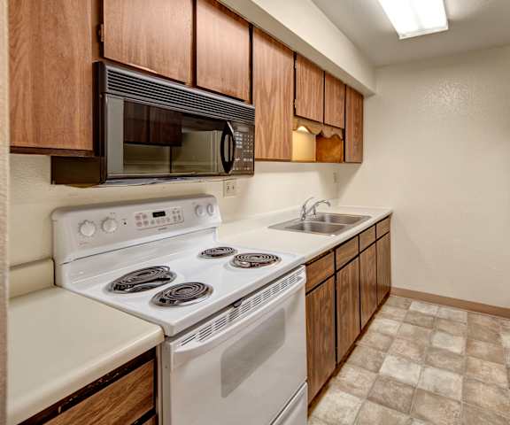 A cozy kitchen with white appliances, kitchen utensils, overhead lighting, light countertops with lots of room, and large wooden cabinets at Terrace on the Lake Apartment Homes. 